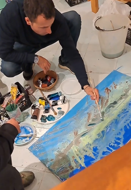 A participant painting a large blue and green abstract landscape on the floor during a creative workshop at Under the Arch Art Gallery.